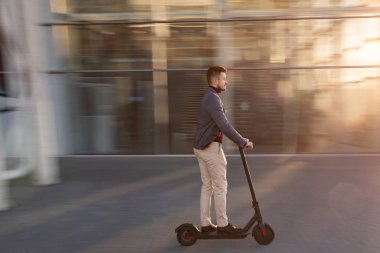Gün batımında havaalanının yanındaki yolda e-scooter kullanan genç yakışıklı bir adam. Modern elektrikli scooter 'la son moda şehir ulaşımı. Çevre dostu hareketlilik kavramı.
