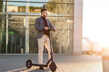 Günbatımında iş merkezinin yanındaki kaldırımda e-scooterlı genç yakışıklı adam. Modern elektrikli scooter 'la son moda şehir ulaşımı. Çevre dostu hareketlilik kavramı.