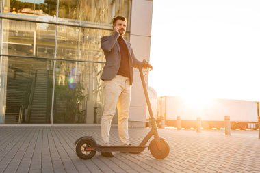 Günbatımında havaalanının yanındaki kaldırımda duran ve akıllı telefondan konuşan, e-scooterlı genç yakışıklı bir adam. Modern elektrikli scooterda trend şehir ulaşımı. Çevre dostu hareketlilik kavramı.