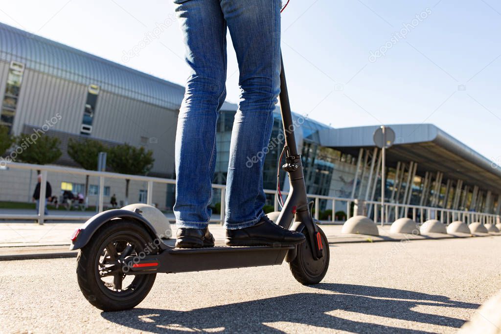 Legs of a man standing on e-scooter parked on sidewalk at cityscape on the sunset. Trendy urban transportation on modern electric scooter. Eco friendly mobility concept.