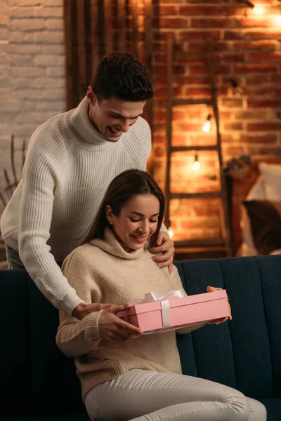 Hombre guapo presentando un regalo a su hermosa novia y sonriendo. Hermosa pareja joven en casa disfrutando de pasar tiempo juntos.Día de San Valentín, las relaciones y el concepto de personas — Foto de Stock