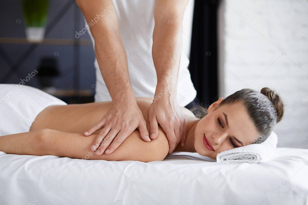 Young beautiful woman enjoying back and shouders massage in spa.Professional massage therapist is treating a female patient in apartment.Relaxation,beauty,body and face treatment concept.Home massage