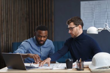 Çok ırklı mühendis ekibi mavi baskılar ve mimar ekipmanlarıyla birlikte çalışıyor, taslak çiziyorlar, tartışıyorlar ve inşaat projesini planlıyorlar. Ortaklar iş yerinde tartışır. İş inşa kavramı