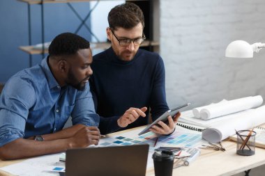 Çok ırklı mühendis ekibi tablet bilgisayarda çalışıyor. Mimarlar bina renklerini renk örnekleri, çizim ve inşaat projeleri kullanarak seçerler. İş yerinde tartışan ortaklar