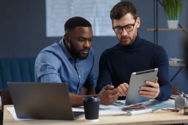 Çok ırklı mühendis ekibi tablet bilgisayarda çalışıyor. Mimarlar bina renklerini renk örnekleri, çizim ve inşaat projeleri kullanarak seçerler. İş yerinde tartışan ortaklar
