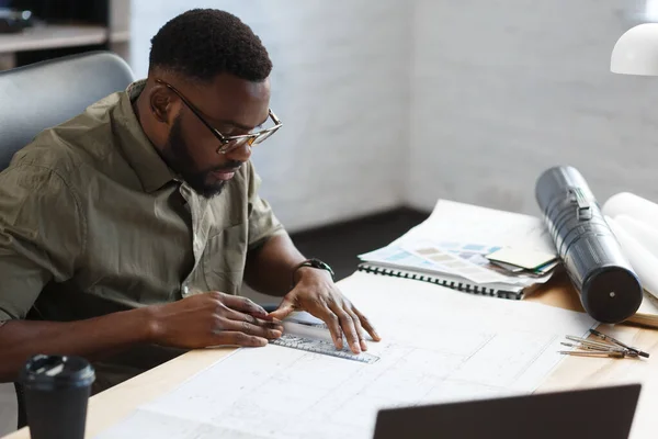 Афро-американский архитектор, работающий в офисе с чертежами. Инженер проверяет архитектурный план, рисует строительный проект. Портрет черного красавца, сидящего на рабочем месте. Концепция бизнеса — стоковое фото