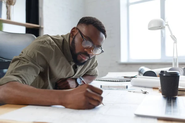 Afro-Amerikaanse architect werkt in het kantoor met blauwdrukken. Ingenieur inspecteren architectonisch plan, schetsen van een bouwproject. Portret van een zwarte knappe man op het werk. Bedrijfsconcept — Stockfoto