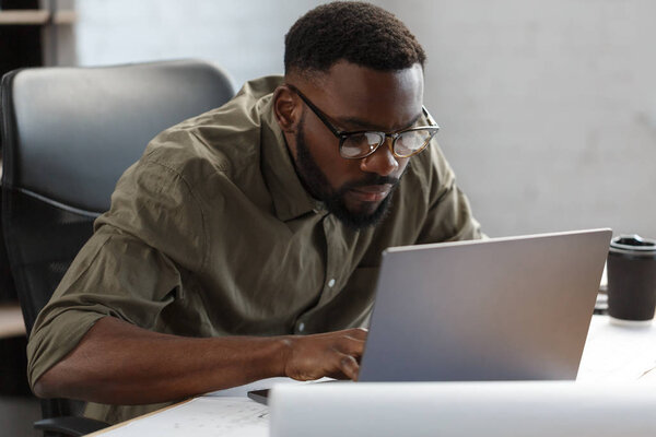 Афро-американский архитектор работает в офисе с laptop.Business портрет черного красивого бородатого мужчину в очках, сидящего на рабочем месте.
