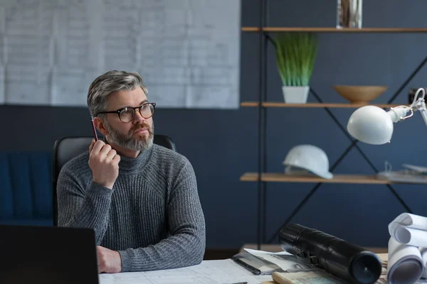 Designer som arbetar i kontor med bärbar dator. Arkitekt funderar över arkitektonisk plan, söker nya idéer för byggprojekt. Porträtt av stilig skäggig man som sitter på arbetsplatsen. Affärsidé — Stockfoto