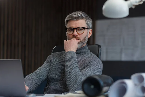 Diseñador trabajando en oficina con laptop. Arquitecto reflexiona sobre el plan arquitectónico, buscando nuevas ideas para el proyecto de construcción. Retrato del guapo barbudo sentado en el lugar de trabajo. Concepto empresarial — Foto de Stock