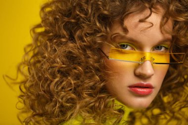 Close up fashion studio portrait of young beautiful women with curly hair. Nice girl with afro hair and bright make up wearing yellow eyeglasses over yellow background. Beauty and hair care concept