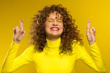 Young beautiful smiling women with curly hair and crossing fingers and wishing good luck. Funny girl with wavy hairstyle and bright make up isolated over yellow background. Yellow lifestyle