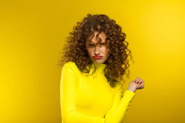 Portrait of young beautiful women with curly hair and suspicious skeptical expression. Nice girl with wavy hairstyle and bright make up isolated over yellow background. Yellow lifestyle. Copy space