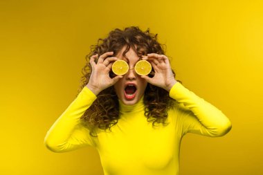 Beautiful curly-haired women with open mouth covering eyes with lemon halves, being surprised and shocked isolated over yellow background. Reaction on unexpected news. Copy space. Expressive faces