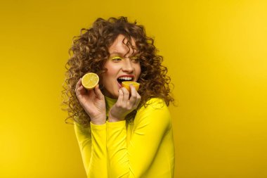 Kıvırcık saçlı, gülümseyen genç ve güzel kadınlar ekşi limon tadarlar. Dalgalı saç stili ve parlak makyajlı komik kız sarı arka planda izole edilmiş. Uzayı kopyala Sarı yaşam tarzı
