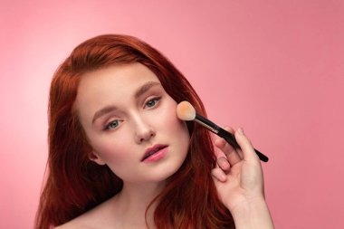 Makyöz pudra sürüyor. Makyaj ustasının eli, genç kızıl saçlı kızın yanaklarını boyuyor. Temiz ciltli ve doğal makyajlı güzel sağlıklı bir kızın portresi. Güzellik Konsepti
