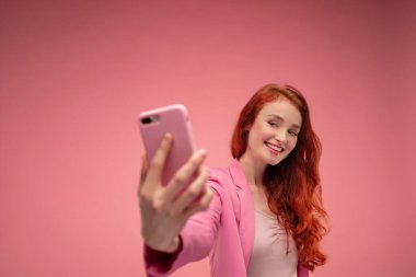 Güzel kızıl saçlı kadın selfie çekiyor. Pembe ceket giyen, elinde pembe akıllı telefon tutan ve pembe arka planda izole edilmiş selfie için poz veren komik kız.