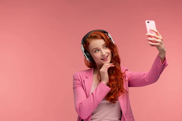 Enjoy listening to music. Beautiful young redhead woman with headphones listening music and making selfie. Funny smiling girl with earphones and mobile phone on pink background — Stock Photo, Image
