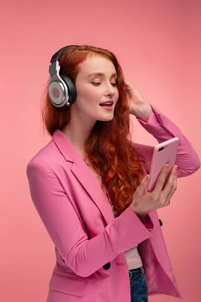 Desfrute de ouvir música. Mulher ruiva bonita com fones de ouvido ouvindo música no telefone inteligente usando o aplicativo de música. Menina sorrindo engraçado com fones de ouvido e telefone celular no fundo rosa — Fotografia de Stock