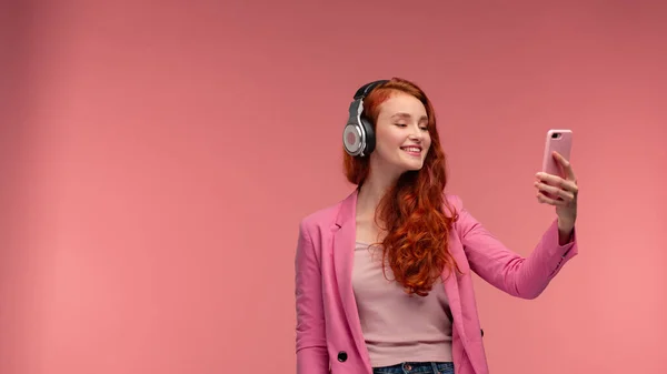 Disfruta escuchando música. Hermosa joven pelirroja con auriculares escuchando música en el teléfono inteligente utilizando la aplicación de música. Chica sonriente divertida con auriculares y teléfono móvil sobre fondo rosa —  Fotos de Stock