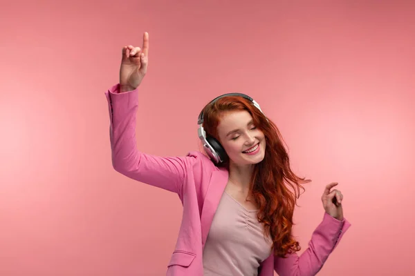 Disfruta escuchando música. Hermosa joven pelirroja en auriculares escuchando música. Chica sonriente divertida en auriculares y chaqueta rosa bailando sobre fondo rosa —  Fotos de Stock
