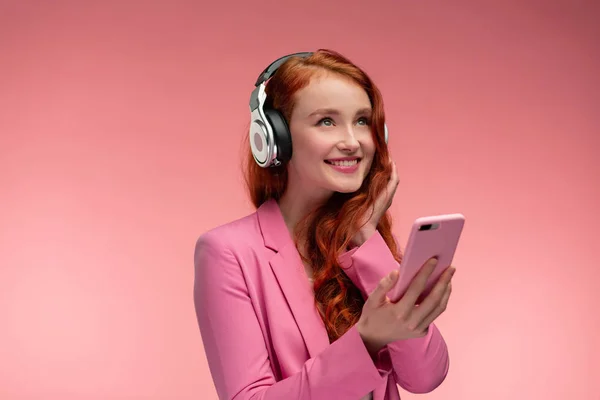 Disfruta escuchando música. Hermosa joven pelirroja con auriculares escuchando música en el teléfono inteligente utilizando la aplicación de música. Chica sonriente divertida con auriculares y teléfono móvil sobre fondo rosa —  Fotos de Stock