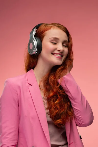 Desfrute de ouvir música. Mulher ruiva bonita em fones de ouvido ouvir música. Menina sorrindo engraçado em fones de ouvido e jaqueta rosa dançando no fundo rosa — Fotografia de Stock