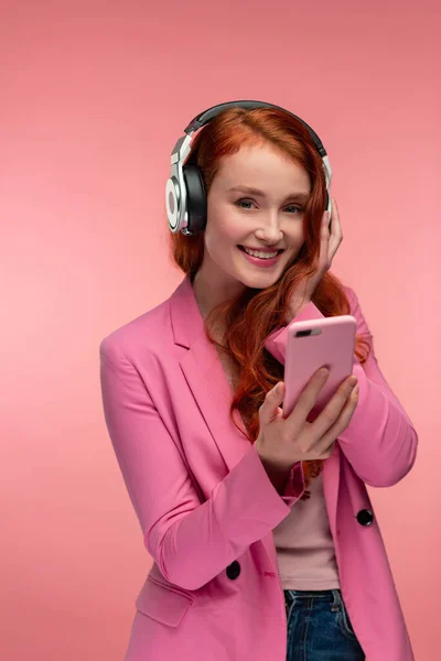 Disfruta escuchando música. Hermosa joven pelirroja con auriculares escuchando música en el teléfono inteligente utilizando la aplicación de música. Chica sonriente divertida con auriculares y teléfono móvil sobre fondo rosa —  Fotos de Stock