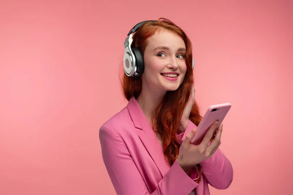 Disfruta escuchando música. Hermosa joven pelirroja con auriculares escuchando música en el teléfono inteligente utilizando la aplicación de música. Chica sonriente divertida con auriculares y teléfono móvil sobre fondo rosa —  Fotos de Stock
