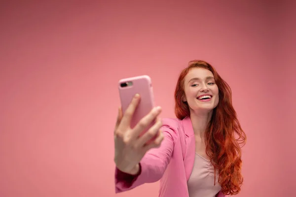 Beautiful young redhead woman making selfie. Funny smiling girl wearing pink jacket holding pink smartphone and posing for selfie isolated on pink background — Stock Photo, Image