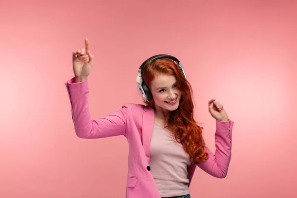 Disfruta escuchando música. Hermosa joven pelirroja en auriculares escuchando música. Chica sonriente divertida en auriculares y chaqueta rosa bailando sobre fondo rosa —  Fotos de Stock