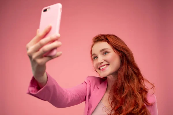 Beautiful young redhead woman making selfie. Funny smiling girl wearing pink jacket holding pink smartphone and posing for selfie isolated on pink background — Stock Photo, Image