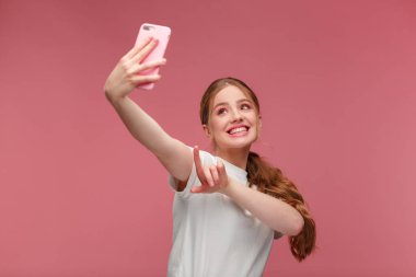 Komik kızıl saçlı kadın selfie çekiyor. Beyaz tişört giyen, elinde pembe akıllı telefon tutan gülümseyen kız, kamerada yüz ifadeleri yapan, pembe arka planda izole edilmiş selfie için poz veren kız.