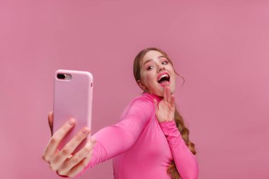 Komik kızıl saçlı kadın selfie çekiyor. Pembe bluz giyen, elinde pembe akıllı telefon tutan gülümseyen kız, kamerada yüz ifadeleri yapan, pembe arka planda izole edilmiş selfie için poz veren kız.