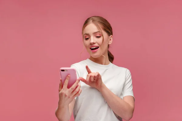 Funny young redhead woman holding pink smartphone and smiling.Happy girl using mobile phone apps, texting message, browsing internet, looking at smartphone. Young people working with mobile devices. — Stock Photo, Image
