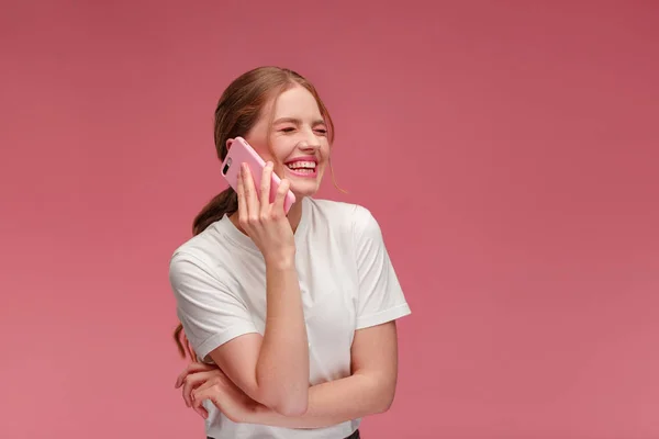 Portrait of cute happy redhead girl wearing white t-shirt talking on mobile phone and smiling isolated over pink background. Copy space. Young people working with mobile devices — Stock Photo, Image