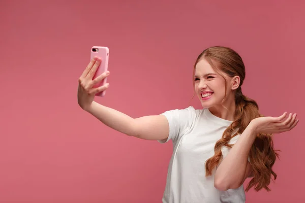 Παράξενη κοκκινομάλλα που βγάζει σέλφι. Χαμογελαστό κορίτσι φορώντας λευκό μπλουζάκι κρατώντας ροζ smartphone, κάνοντας γκριμάτσες στην κάμερα, ποζάροντας για selfie απομονωμένη σε ροζ φόντο — Φωτογραφία Αρχείου