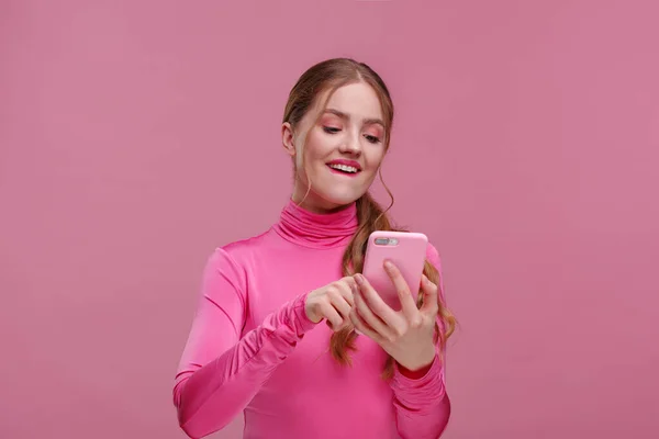 Найкращий день у світі. Здивована молода руда жінка тримає рожевий смартфон, посміхаючись і висловлюючи позитивізм. Щаслива дівчина отримала шокуючі позитивні новини. Копіювати простір. Молоді люди, що працюють з мобільними пристроями — стокове фото