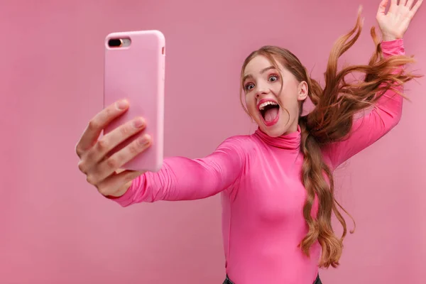 Joven pelirroja divertida haciendo selfie. Chica sonriente con blusa rosa sosteniendo teléfono inteligente rosa, haciendo caras en la cámara, posando para selfie aislado sobre fondo rosa — Foto de Stock