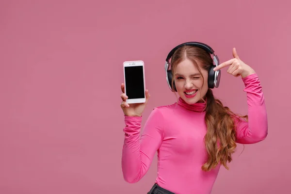 Menina ruiva engraçado mostrando telefone celular tela em branco na câmera. Jovem sorrindo e piscando mulher usando fones de ouvido demonstrando exibição de smartphone isolado sobre fundo rosa. Espaço de cópia — Fotografia de Stock