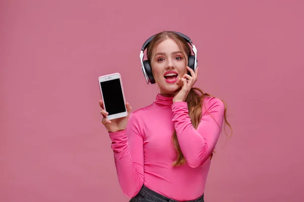 Menina ruiva engraçado mostrando telefone celular tela em branco na câmera. Mulher sorrindo jovem usando fones de ouvido demonstrando exibição de smartphone isolado sobre fundo rosa. Espaço para cópia. App para o seu negócio . — Fotografia de Stock