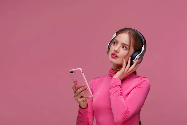 Enjoy listening to music. Beautiful young redhead woman with headphones listening music on smart phone using music app. Funny smiling girl with earphones and mobile phone on pink background