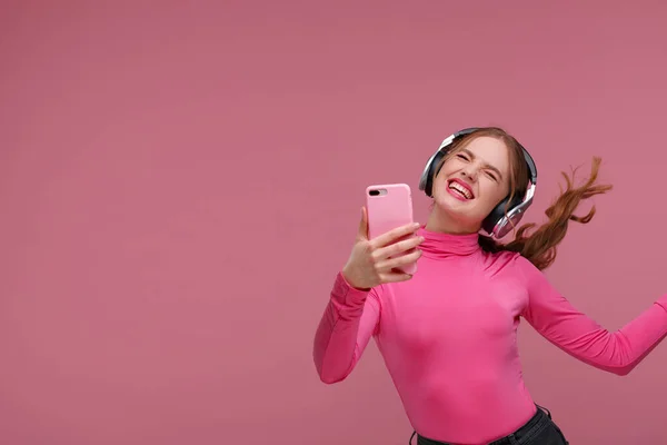 Enjoy listening to music. Beautiful young redhead woman with headphones listening music and making selfie. Funny emotional smiling girl with earphones and mobile phone on pink background