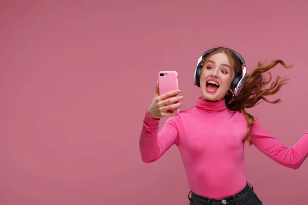 Disfruta escuchando música. Hermosa joven pelirroja con auriculares escuchando música y haciendo selfie. Divertida chica sonriente emocional con auriculares y teléfono móvil sobre fondo rosa —  Fotos de Stock