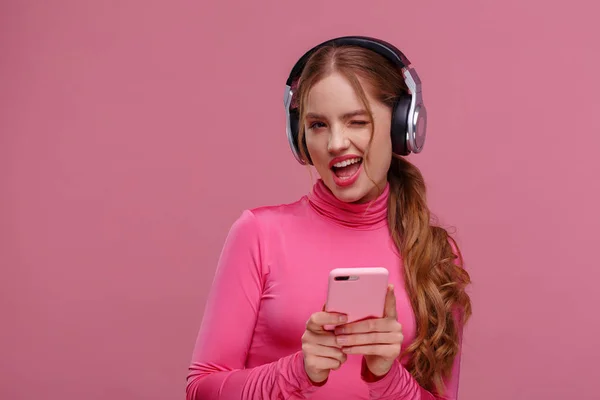 Menina ruiva engraçado segurando telefone celular à espera de mensagens e parabéns de amigos. Jovem mulher sorridente usando fones de ouvido segurando smartphone sobre fundo rosa. Espaço de cópia — Fotografia de Stock