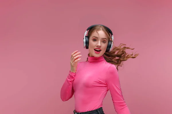 Geniet van muziek luisteren. Mooie jonge roodharige vrouw in koptelefoon die muziek luistert. Grappig lachend meisje in oortjes en roze blouse dansend op roze achtergrond. Ontspanning en stressbeheersing — Stockfoto