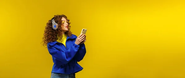 Enjoy listening to music. Beautiful young curly-haired woman with headphones listening music,singing and dancing. Funny emotional smiling girl with earphones and mobile phone on yellow background — Stock Photo, Image