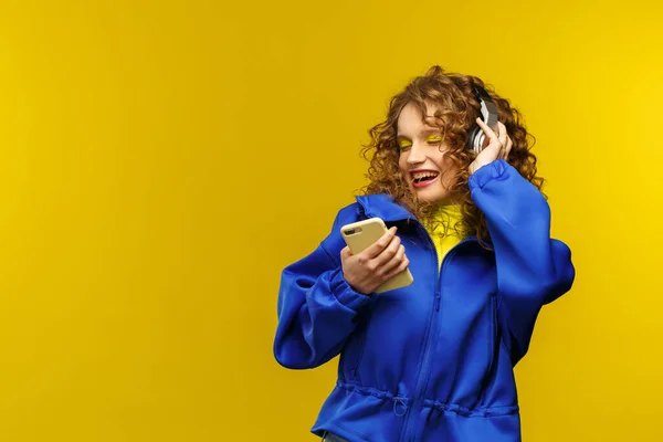 Enjoy listening to music. Beautiful young curly-haired woman with headphones listening music,singing and dancing. Funny emotional smiling girl with earphones and mobile phone on yellow background — Stock Photo, Image