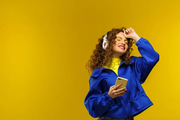 Enjoy listening to music. Beautiful young curly-haired woman with headphones listening music,singing and dancing. Funny emotional smiling girl with earphones and mobile phone on yellow background — Stock Photo, Image