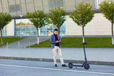 Akıllı telefonu tutan yakışıklı genç adam gün batımında havaalanı yakınlarında bir e-scooter kiralamaya hazırlanıyor. Modern elektrikli scooter 'la son moda şehir ulaşımı. Çevre dostu hareketlilik kavramı